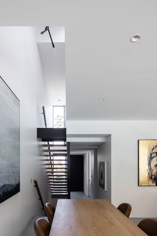 A bright contemporary dining area viewed toward an open stairway and hallway. The room has white walls and ceiling, recessed downlights, a large light timber dining table with brown leather dining chairs, oversized abstract artwork, and a dramatic open-riser staircase with dark structural elements and a skylit or double-height void.