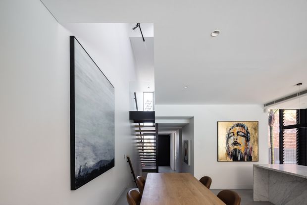A bright contemporary open-plan interior showing a dining area with a long timber dining table and dark upholstered chairs, adjacent to a stone-clad kitchen island. White walls and ceilings, black-framed glazing with blinds, recessed lighting, a large monochrome artwork, and an open stairway with horizontal timber treads create a clean, refined setting.