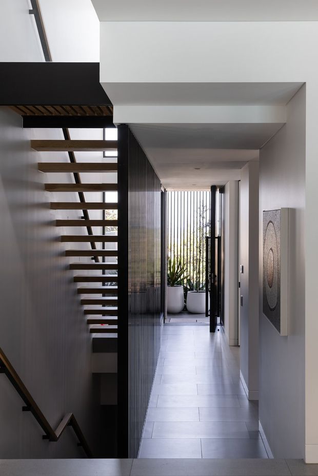 A contemporary interior hallway viewed beside an open-riser staircase. The corridor has pale tiled flooring, white walls and ceiling, a dark vertical slatted screen, and black-framed glazing leading toward a bright planted outdoor area. Floating timber stair treads and dark handrails create a strong linear architectural feature, while a monochrome wall artwork adds a subtle decorative element.