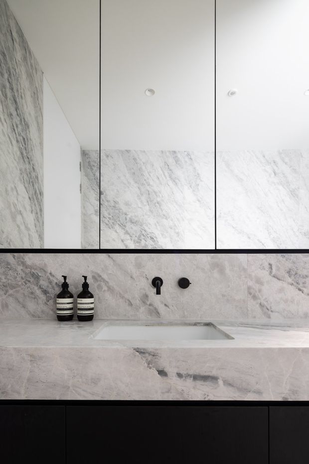 Close-up view of a refined contemporary bathroom vanity. A pale grey-white marble or stone surface extends across the benchtop and backsplash, with an integrated rectangular basin, matte black wall-mounted tapware, black lower cabinetry, and mirrored wall cabinets above. The restrained monochrome palette and clean lines create a minimalist, high-end appearance.