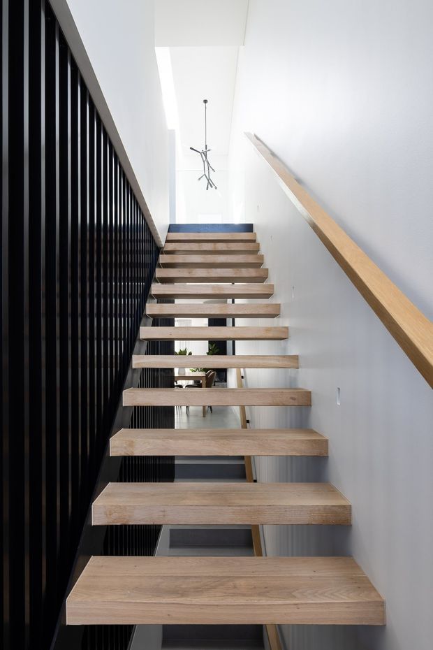 A bright, narrow interior stairway with floating light timber treads ascending between a white wall and a tall black vertical-slat balustrade. A matching timber handrail runs along the right wall, while a minimalist sculptural pendant hangs above the stair void. A dining or living area is partially visible beneath and beyond the stairs.