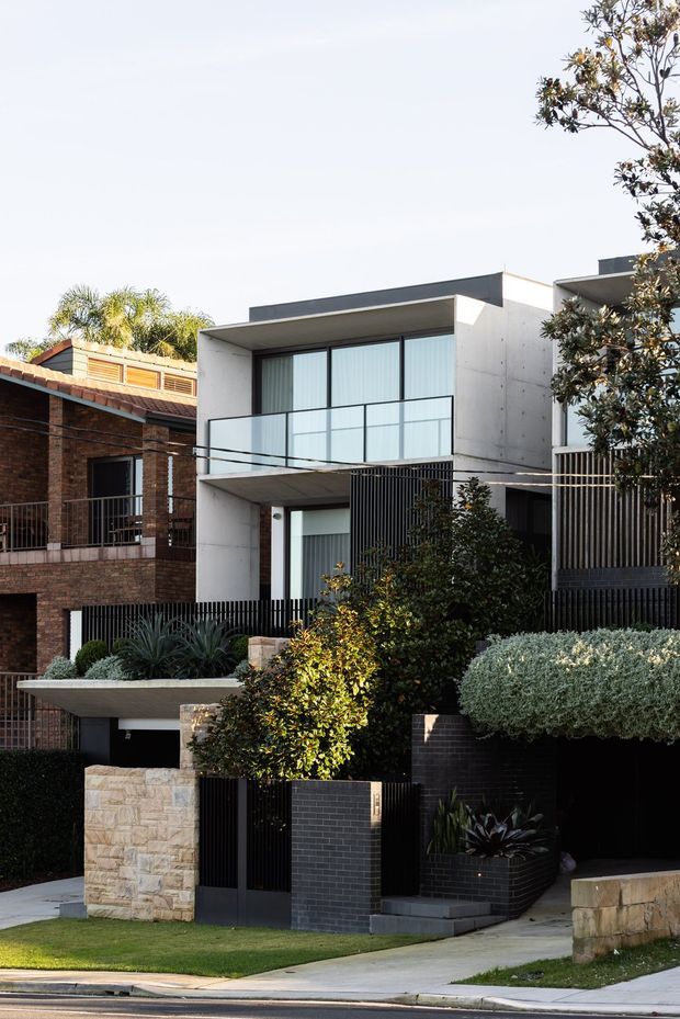Street-facing photograph of a contemporary multi-storey urban residence with a rectilinear white rendered facade, dark charcoal roofline and brickwork, broad glazed openings, glass balcony balustrade, and black vertical metal screening. The property includes layered landscaped front gardens, rendered and dark brick retaining walls, stone-clad boundary elements, and an integrated garage or covered parking area. A neighbouring traditional brick home is visible to the left.