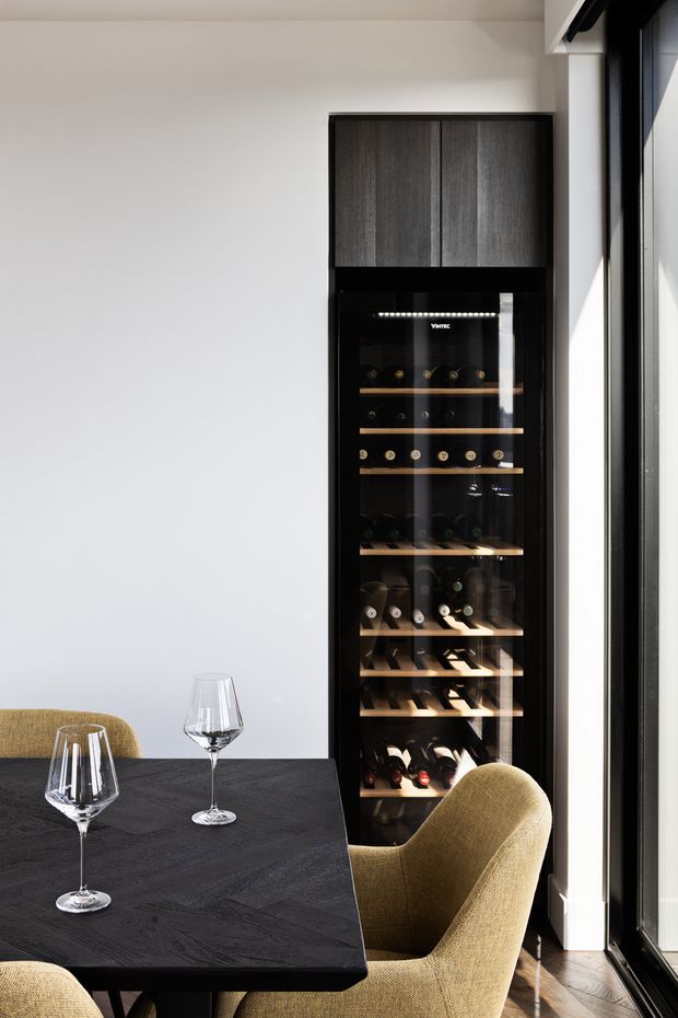 A refined dining area with a dark rectangular dining table, two wine glasses, and upholstered mustard-toned dining chairs. A tall built-in glass-front wine refrigerator with dark wood cabinetry above is set beside a large black-framed window or glazed door. The room has white walls, timber flooring, and strong natural light, creating a contemporary, sophisticated interior.