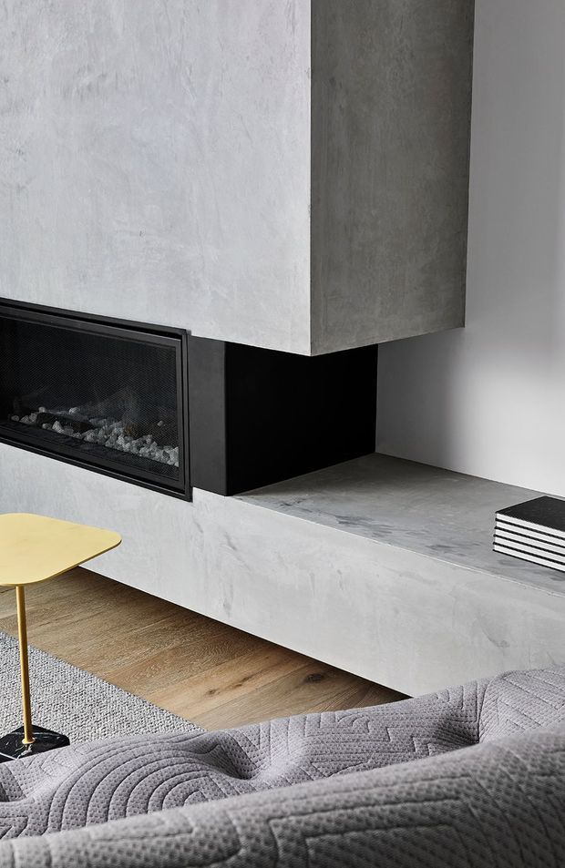 A close-up contemporary living room vignette centred on a built-in linear fireplace with a black metal surround, set within a sculptural grey plaster or concrete media wall and bench. Light timber flooring, a pale textured rug, a small mustard-yellow side table, and a grey quilted sofa create a restrained, modern interior palette.