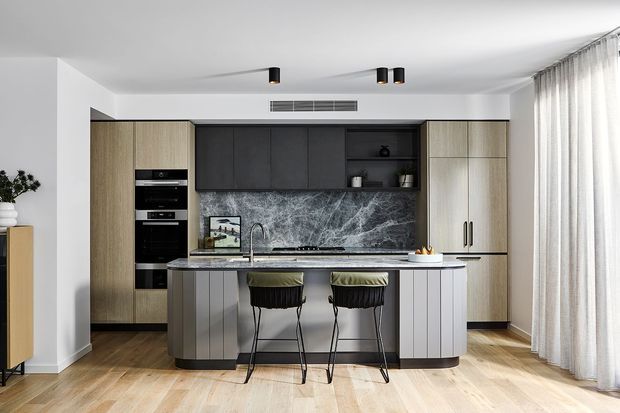 Contemporary kitchen with a central island and two bar stools, framed by tall timber-look cabinetry and dark overhead cabinets. A dramatic grey marble-look splashback, integrated appliances, pale timber flooring, white walls, and sheer full-height curtains create a refined, warm-neutral interior.
