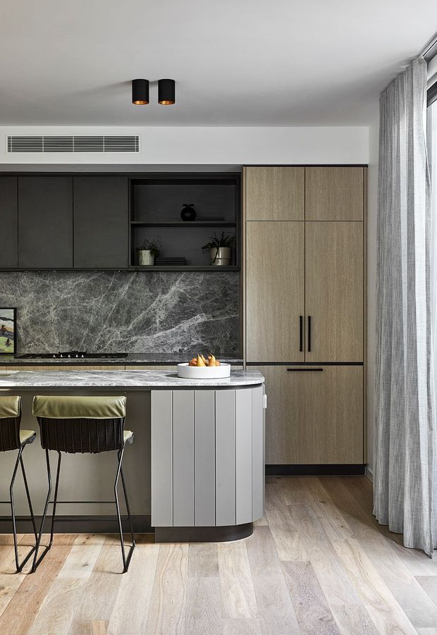 A refined contemporary kitchen with a compact curved island and two bar stools. The space features dark charcoal cabinetry, a dramatic grey marble-look splashback, light timber-grain pantry cabinetry, pale timber flooring, and full-height sheer curtains beside a large window. Black cylindrical ceiling lights and dark hardware add contrast to the neutral palette.