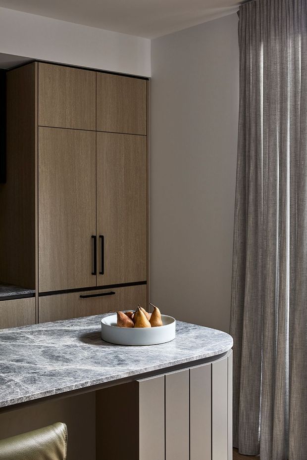 A refined contemporary kitchen interior featuring a compact island with a dark veined stone benchtop and vertically fluted light cabinetry. Full-height wood-grain storage cabinetry with black handles lines the rear wall, while floor-to-ceiling textured grey curtains frame the right side. A bowl of pears sits on the island, and a partial upholstered bar stool is visible in the foreground.