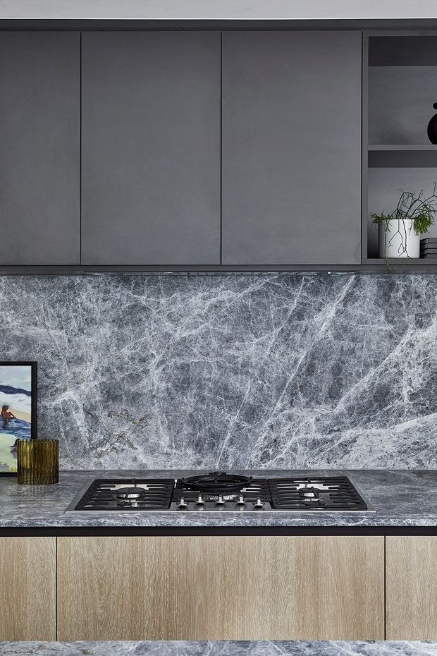 Close-up view of a contemporary kitchen featuring a dramatic grey veined stone splashback and benchtop, dark grey flat-panel overhead cabinetry, light timber-grain lower cabinetry, and a built-in black gas cooktop. Open shelving with a small plant is visible at the right.