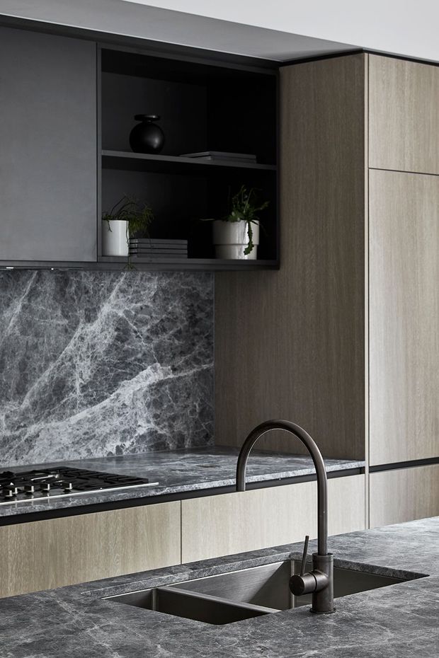 Close-up view of a refined contemporary kitchen featuring dark grey marble-look stone benchtops and splashback, light timber-grain cabinetry, matte black open shelving and a black gooseneck mixer tap. A gas cooktop, undermount double sink, potted plants and restrained dark accessories complete the composition.