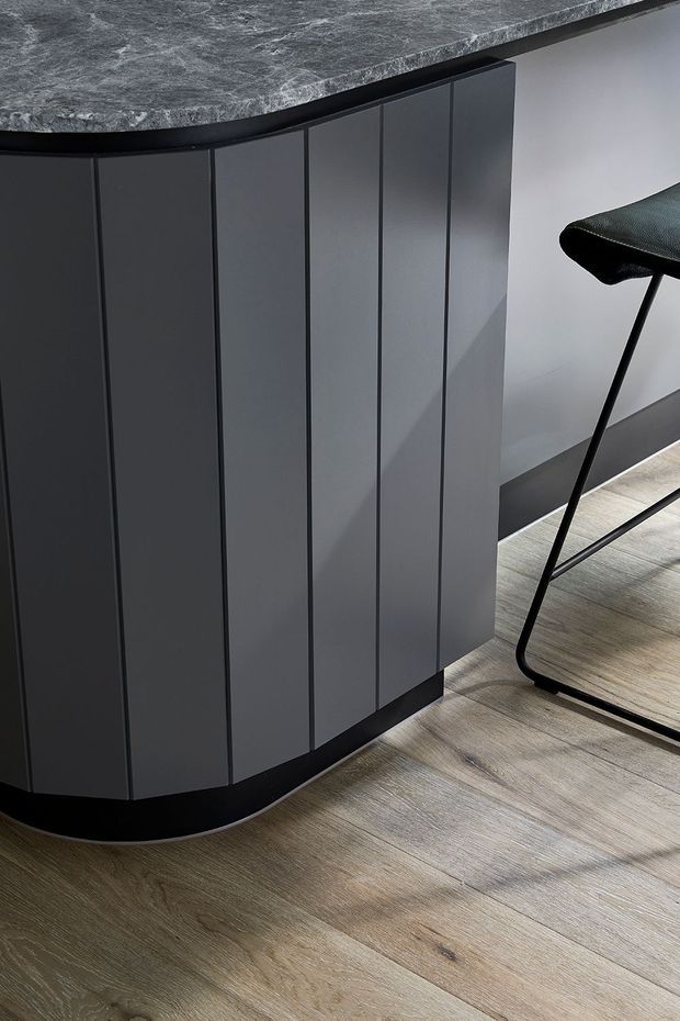 Close-up of a contemporary curved kitchen island or built-in cabinet with dark charcoal vertical-panel cabinetry and a mottled dark stone benchtop. The island has a rounded end and recessed dark plinth, set against a light wall with pale timber-look flooring. A black metal bar stool is partially visible at right.
