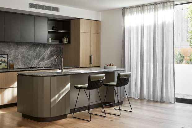 A bright contemporary kitchen with a large island and two bar stools, featuring dark cabinetry, warm timber-toned tall storage, a dramatic grey marble-look splashback, and light timber flooring. Full-height sheer curtains open to a small outdoor terrace, bringing in abundant natural light.