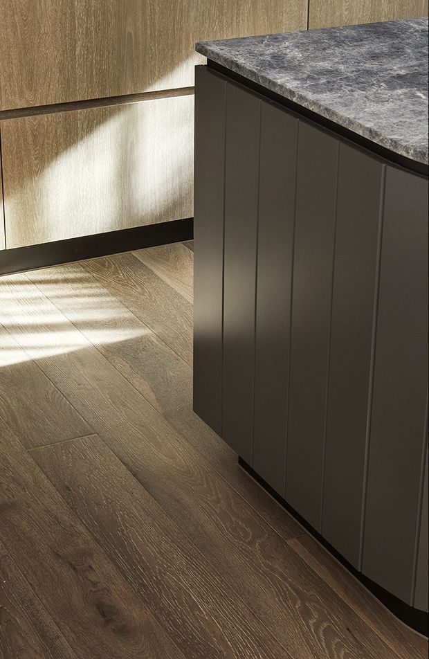 Close-up view of a contemporary kitchen island with dark vertically panelled cabinetry and a dark grey stone-look benchtop. Warm wood-toned cabinetry and matching timber flooring create a refined, earthy interior, with sunlight casting patterned shadows across the floor.