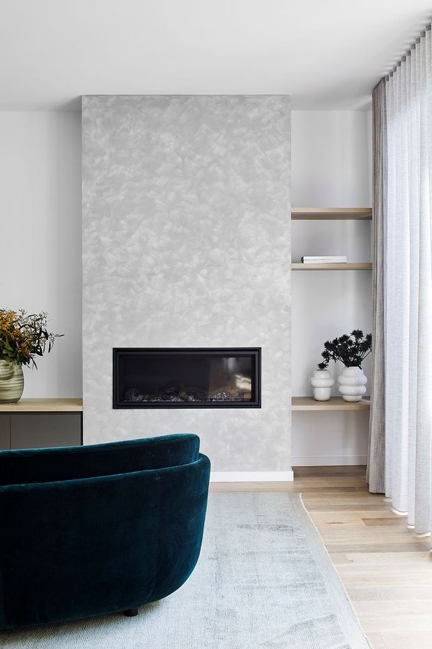 A refined living room with a textured pale feature wall and built-in linear fireplace, flanked by open timber shelves and cabinetry. A deep teal velvet sofa, pale rug, light timber flooring, sheer full-height curtains, and restrained decorative styling create a calm contemporary interior.