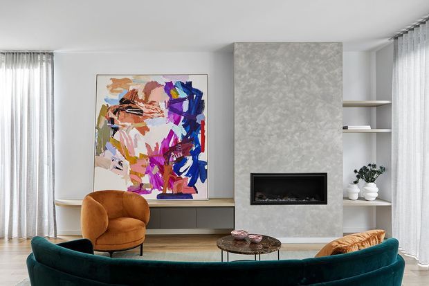 Contemporary living room with a deep teal curved sofa, rust-toned lounge chair, round coffee table, abstract colourful artwork, built-in low cabinetry, open shelving, and a recessed fireplace set into a textured grey feature wall. Full-height sheer curtains frame the room, with warm timber flooring and neutral finishes creating a refined, inviting interior.