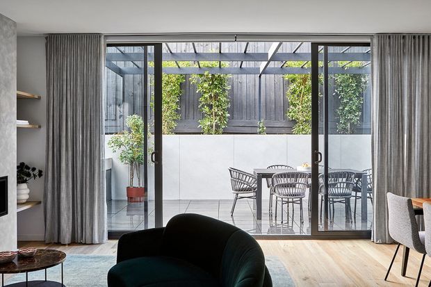 A contemporary living room opens through full-height sliding glass doors to a private paved courtyard with outdoor dining furniture, a pergola-like overhead structure, raised planters and dark timber fencing. The interior features pale timber flooring, full-height grey curtains, a dark curved lounge chair, built-in shelving and neutral furnishings.