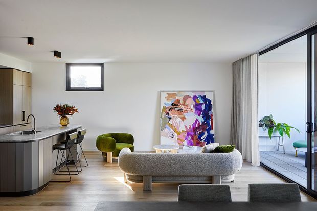 Bright open-plan contemporary living area with a compact kitchen at left, a curved island with bar stools, a textured sofa facing a colourful abstract artwork, and a dining table in the foreground. Full-height glazed doors with sheer curtains open toward a balcony or terrace, bringing in abundant natural light. The space features pale timber flooring, white walls and ceiling, dark-framed glazing, and restrained modern detailing.