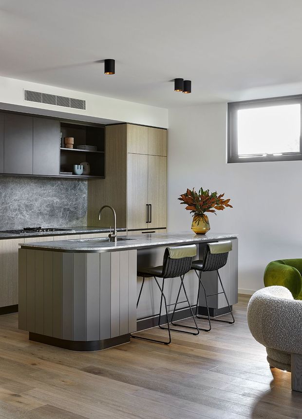 A contemporary open-plan kitchen with a large island, integrated cabinetry, stone benchtops and splashback, timber-look flooring, and two black bar stools. The kitchen combines dark grey and warm wood cabinetry with a bright white ceiling and walls. A partially visible living area with green and cream lounge chairs appears at the right edge.