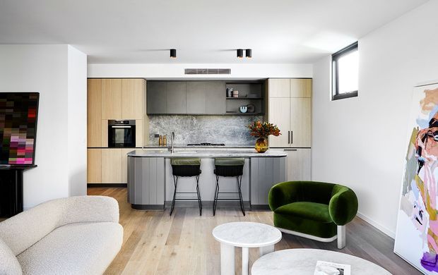 Open-plan contemporary kitchen and living area with pale timber cabinetry, a dark grey island, stone-look splashback, timber flooring, and a small dining or lounge setting. The room is bright and minimally styled, with a green velvet accent chair and artwork adding colour.