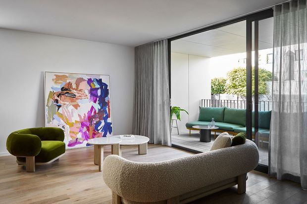 Contemporary living room with warm timber flooring, white walls, sculptural boucle seating, a green armchair and sofa, a colorful abstract artwork, and full-height sliding glass doors opening to a balcony or terrace. Sheer grey curtains soften the bright indoor-outdoor connection.