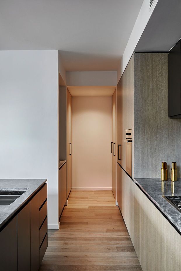 A narrow contemporary kitchen galley with integrated timber-grain cabinetry on both sides, dark stone benchtops, and warm timber flooring. The view looks down a softly lit passage toward the rear of the kitchen, with built-in appliances, a sink, cooktop, and subtle brass accessories.