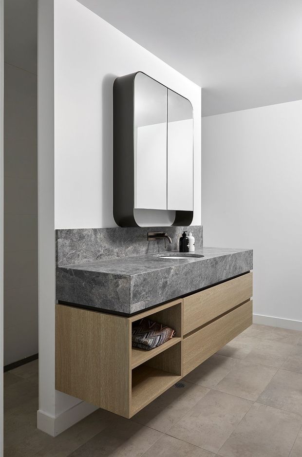 Contemporary bathroom vanity featuring a floating light timber cabinet with open shelving and drawers, a thick grey veined stone benchtop with integrated basin, wall-mounted tapware, and a rounded rectangular black-framed mirror. The space has white walls, grey floor tiles, and a clean minimalist composition.
