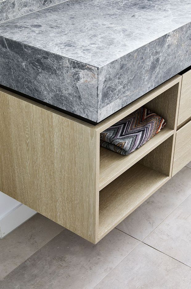 Close-up detail of a built-in light timber-grain cabinet with open shelving and drawers, topped by a thick grey veined stone benchtop. Large pale grey floor tiles are visible below, with a folded patterned textile stored on the shelf.