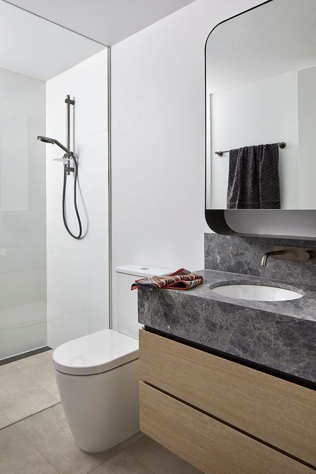 Contemporary bathroom featuring a walk-in shower with dark metal fixtures, a wall-hung toilet, and a floating timber-look vanity with a grey stone benchtop and integrated oval basin. A large rounded-corner mirror, monochromatic wall tiles, and neutral flooring create a clean, refined fit-out.