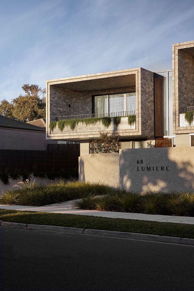 Street-facing view of a contemporary multi-unit residential building with projecting box-like balconies and deep rectangular openings. The facade uses textured tan-brown masonry, with timber-toned vertical cladding and large glazed doors/windows. A pale rendered entry wall marked “68 LUMIERE” fronts a landscaped verge with ornamental grasses and low planting.