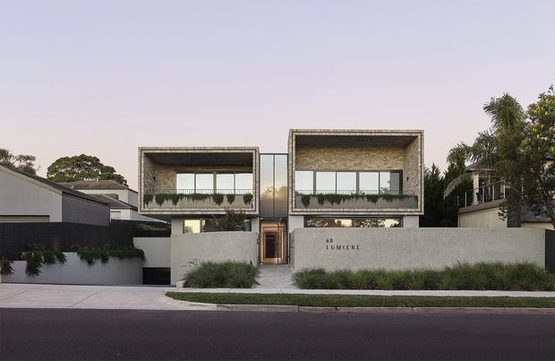 Street-facing view of a contemporary multi-storey residential building with two prominent box-like upper volumes, textured masonry or concrete facades, extensive horizontal glazing, recessed balconies, a central entry, and a low rendered boundary wall with landscaped planting.