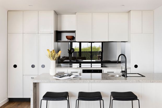 Bright contemporary kitchen with extensive white flat-panel cabinetry, a large pale stone island with integrated seating, black-framed stools, dark fixtures, and a reflective black splashback. A window and glazed doors behind the kitchen provide views to greenery and a balcony or outdoor area.