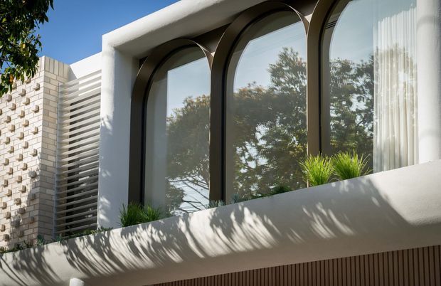 Close-up exterior view of a contemporary building facade featuring a projecting white rendered planter or balcony edge, three tall arched windows with dark metal frames, and textured light masonry with horizontal screening to the left. Reflections of trees are visible in the glazing, with sculptural grasses planted along the ledge.