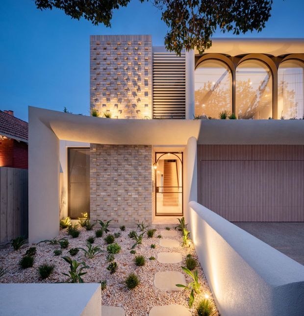 Twilight exterior view of a contemporary multi-level residence with sculptural curved white rendered walls, a pale brick feature wall, arched upper-level glazing, horizontal louvres, and a ribbed garage door. A landscaped gravel forecourt with stepping stones, low grasses, and subtle garden lighting leads to the entrance.
