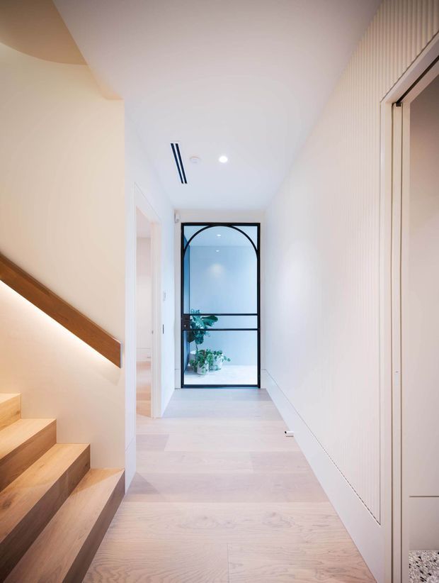 A bright, minimalist residential hallway with pale timber flooring and a matching timber staircase at left. White walls and concealed cabinetry create a clean, streamlined interior, while a black-framed glazed door with a curved arch detail forms a feature at the end of the corridor. A slim linear ceiling vent and recessed downlight add to the contemporary fit-out, with potted greenery visible beyond the glazed door.