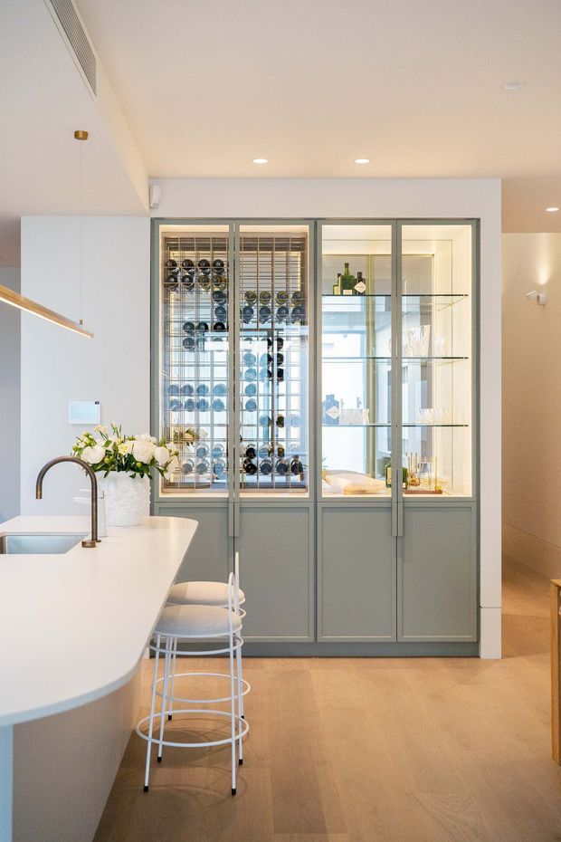 Bright contemporary kitchen featuring a large white waterfall-edge island with an undermount sink and curved brushed-metal tap, paired with white bar stools. A full-height grey-framed glazed display cabinet and integrated wine storage form the focal wall, with glass shelving and bottles visible inside. Pale timber flooring, white walls, recessed ceiling lights, and understated grey cabinetry create a clean, refined interior.