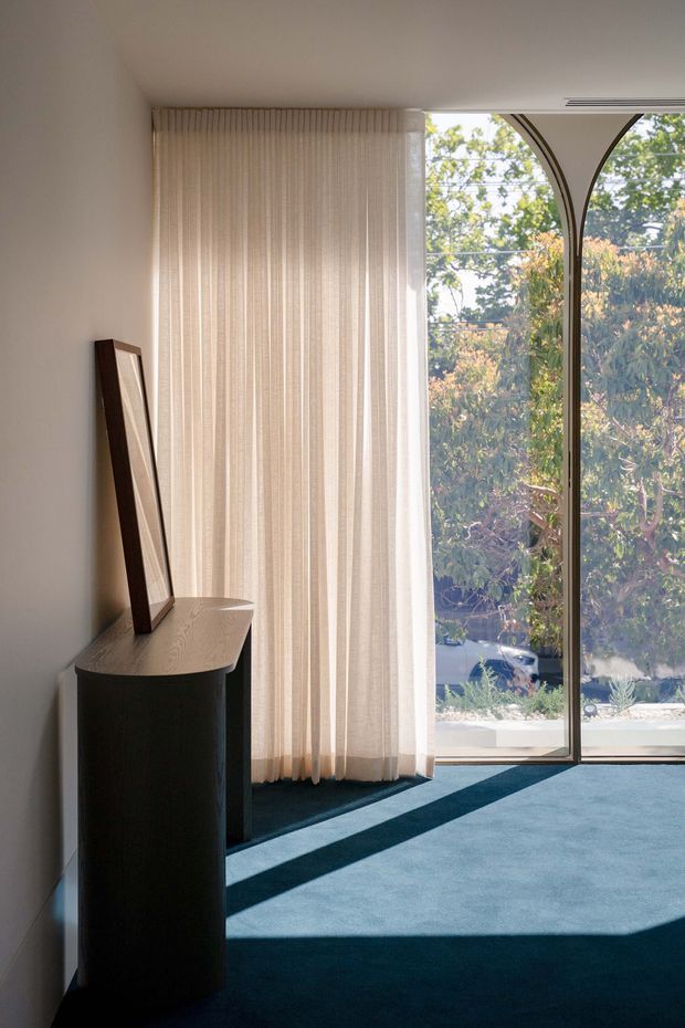Sunlit minimalist interior corner with a dark curved timber console and leaning mirror against a pale wall. Full-height sheer curtains frame a large arched-top glazed opening, while blue carpet and strong daylight shadows define the floor. Trees and part of the exterior are visible through the glass.