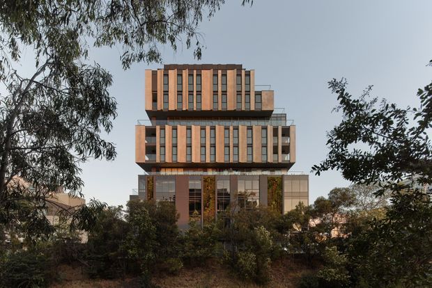 A multi-storey contemporary building viewed externally through mature trees and landscaped vegetation. The facade uses repeated vertical window bays framed by warm terracotta or brick-toned panels, with projecting and recessed floor plates, glass balustrades, and a stepped upper massing. The image primarily presents the building exterior in a suburban or landscaped setting.
