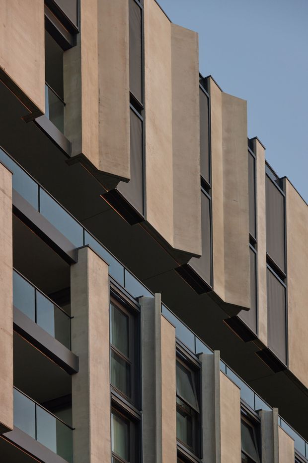 Vertical close-up of a contemporary multi-storey building facade with repeating projecting concrete or rendered wall fins, dark metal-framed windows, recessed balconies, and strong geometric horizontal and vertical lines. The warm beige facade contrasts with dark charcoal structural elements and glazing against a clear blue sky.