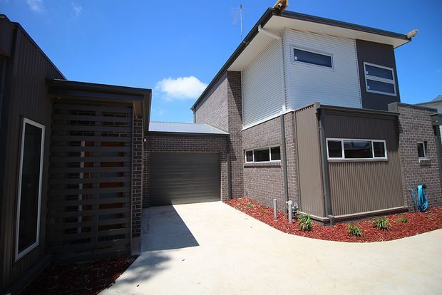 Exterior view of a contemporary multi-level residential building with dark brick and vertical cladding, a projecting upper-storey volume, horizontal windows, an attached garage, and a pale concrete driveway. Red mulch landscaping and a covered timber-screened entry are visible in the foreground.