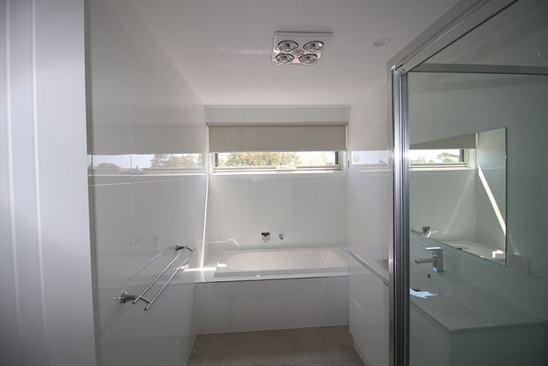 Bright, compact modern bathroom with glossy white wall tiling, a built-in bathtub beneath a horizontal window, wall-mounted towel rails, recessed ceiling lighting, and a glass-enclosed shower or wet-area enclosure at right. A large mirror reflects the vanity and shower area.