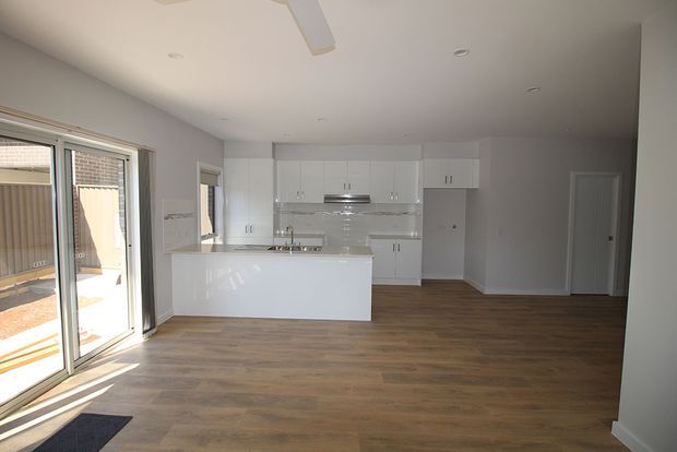 Bright open-plan residential interior with a modern white kitchen, large central island, timber-look flooring, recessed lighting, and sliding glass doors opening to an outdoor area.