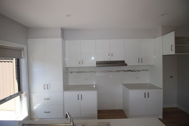 A compact, bright contemporary kitchen with white full-height cabinetry, upper cupboards, a stainless-steel rangehood, pale tiled splashback, light benchtops, and timber-toned flooring. A window provides natural light, and a sink with a curved tap is visible in the foreground.