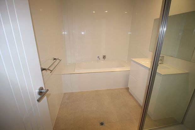 Bright, modern bathroom viewed through an open door, featuring a built-in bathtub with tiled surround, a glass-enclosed shower, white vanity cabinetry, large wall mirror, chrome fixtures, glossy white wall surfaces, and beige floor tiles.