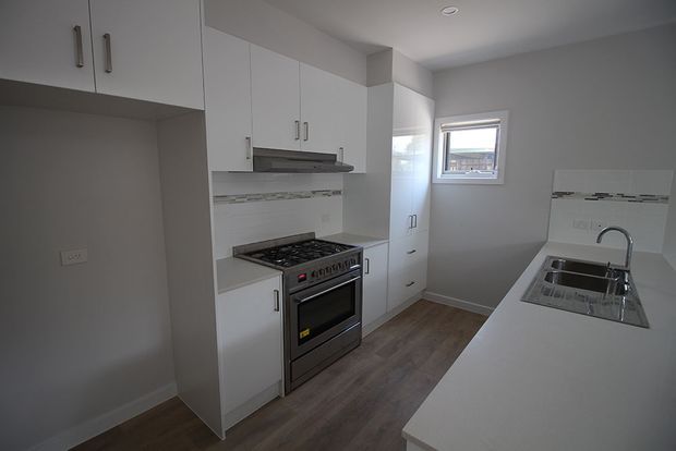 Compact modern kitchen with white flat-panel cabinetry, a freestanding stainless-steel oven and cooktop, grey benchtops, a narrow tiled splashback, timber-look flooring, and a white island with an integrated stainless-steel sink beneath a small window.