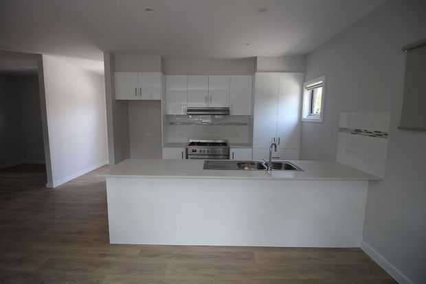 A bright, compact contemporary kitchen with white full-height cabinetry, a large plain-front island with integrated sink and chrome tapware, stainless-steel appliances, and a subtle tiled splashback. Warm timber-look flooring contrasts with the white cabinetry and light grey walls. An adjoining open area is visible to the left.