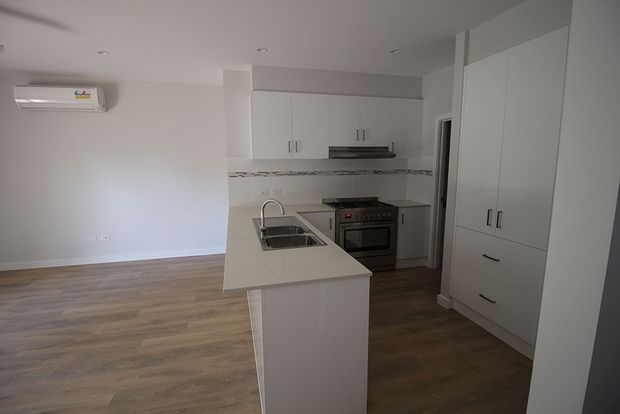 Bright, compact open-plan kitchen with a white island featuring an inset sink, white overhead and full-height cabinetry, integrated storage, stainless-steel appliances, and a narrow decorative tiled splashback. The kitchen opens to a simple living area with warm timber-look flooring and white walls.