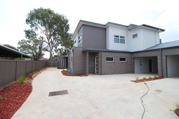 Exterior view of a contemporary multi-level residential building, featuring a rendered or painted brick lower level, light vertical cladding above, a dark metal roof, and a broad concrete driveway or courtyard. A covered carport/garage area and landscaped garden beds are visible.