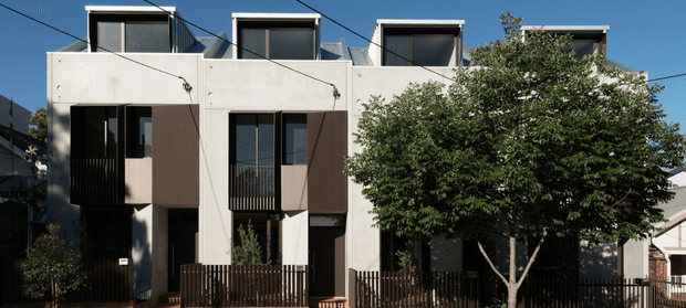 Street-facing view of a contemporary multi-storey residential building composed of several narrow attached units. The facade uses pale rendered panels with dark vertical window and balcony elements, projecting box-like forms, rooftop clerestory or pop-up windows, timber-toned privacy fencing, and a flat roofline. Mature street trees partially screen the lower facade.