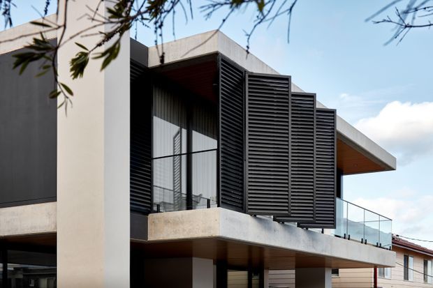 Contemporary multi-level residential facade with projecting concrete balconies, dark horizontal louvred screens, extensive glazing, warm timber soffits, and frameless glass balustrades. The view focuses on the building exterior, with neighbouring structures and sky in the background.
