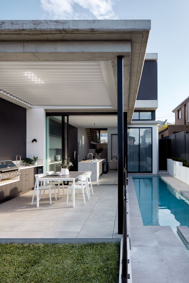 A contemporary residential courtyard viewed from outdoors, featuring a covered outdoor kitchen and dining area opening directly into a bright interior kitchen/living space. Large-format grey paving, dark-framed glazing, white outdoor furniture, a narrow rectangular swimming pool, and strong horizontal concrete roof forms define the scene.