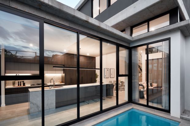 Contemporary courtyard-facing kitchen and living area with full-height black-framed glazing, exposed concrete beams and walls, dark timber cabinetry, a pale stone island, and a turquoise swimming pool in the foreground. The image looks into a refined open-plan interior from the courtyard, with a glimpse of stairs and living furnishings beyond.